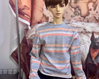 May include: A doll wearing a gray long-sleeve shirt with horizontal stripes in orange and white. The doll is also wearing black pants. A decorative dog statue is in the background.