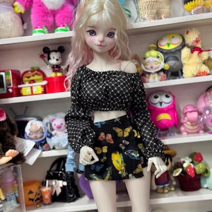 May include: A doll with blonde hair and pink highlights, wearing a black and white checkered crop top and a black skirt with a butterfly pattern. The doll is also wearing pink socks. Shelves filled with various toys and figurines are in the background.