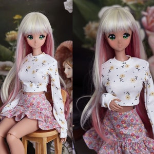 May include: A doll with long blonde and pink hair wearing a white crop top with a floral pattern and a pink and white floral skirt.