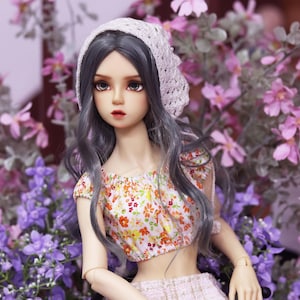 May include: A doll with long gray hair wearing a white crocheted hat, a floral print crop top, and a white skirt. The doll is sitting in front of a background of purple flowers.