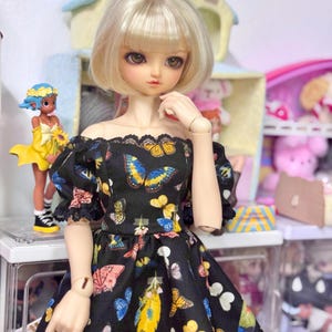 May include: A doll with a blonde bob hairstyle wearing a black off-the-shoulder dress adorned with colorful butterfly patterns. The doll is posed in front of a shelf with other dolls and decorative items.