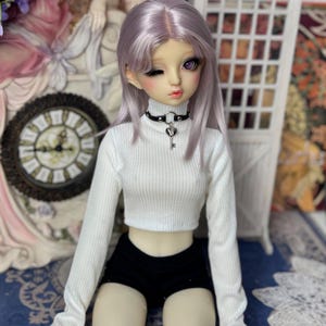 May include: A fashion doll with lavender hair and purple eyes, wearing a white ribbed turtleneck crop top, black shorts, and a black choker with a heart and key pendant. The doll is posed in a seated position.