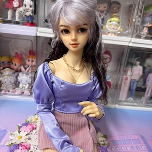 May include: A doll with long, wavy, silver and black hair wearing a lavender blouse with puffed sleeves and a pink skirt. The doll is sitting on a white crocheted doily with floral designs.