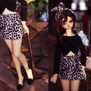 May include: A doll wearing a black long-sleeved top and a mini skirt with a black and white animal print. The doll also has a matching headband. The doll is wearing black shoes.