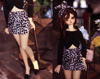 May include: A doll wearing a black long-sleeved top and a mini skirt with a black and white animal print. The doll also has a matching headband. The doll is wearing black shoes.