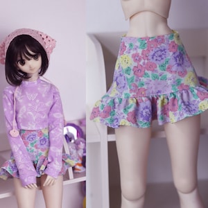 May include: A doll wearing a lavender outfit with a floral skirt and a matching long-sleeved top. The top features a butterfly design. The doll also wears a pink crocheted bonnet. The skirt has a ruffled hem.