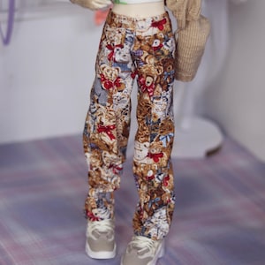 May include: A doll wearing a white t-shirt with a green dinosaur print, a brown cardigan, and brown and beige pants with a teddy bear print. The doll is wearing white sneakers.