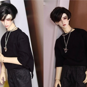May include: A doll with black hair wearing a black t-shirt and black trousers. The doll is wearing two silver necklaces with pendants.