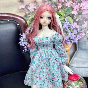 以下が含まれることがあります： ピンクの髪の毛と青い花柄のドレスを着た人形。ドレスは白いレースのトリムとパフスリーブが付いています。人形は黒い椅子に座っており、赤い蓋とJelly Bellyのロゴが付いた透明なガムボールマシンがあります。