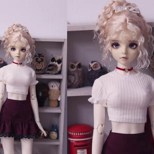 May include: A doll with blonde curly hair wearing a white crop top and a burgundy skirt. The doll is standing in front of a shelf with various decorative items.
