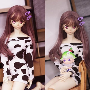 May include: Two dolls with long brown hair wearing cow print dresses. One doll is holding a small green and white doll with a red hat.