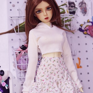 May include: A doll wearing a white ribbed knit hoodie and a floral print skirt with a ruffled hem. The doll has long brown hair and a beaded headband. The background includes a white pegboard and decorative items.