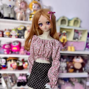 May include: A doll with long auburn hair and purple eyes, wearing a pink floral crop top with long sleeves and a black and white checkered skirt. A pink bow adorns the doll's hair. The doll is posed in front of a shelf filled with various toys.