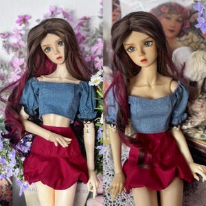 May include: A doll with long brown hair with red highlights, wearing a blue denim crop top and a red skirt. The doll has green eyes and is posed in front of a floral background. The doll's outfit is a two-piece set.