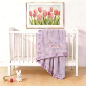 Puede incluir: Una cuna blanca con una manta lavanda bordada con el nombre "Bella" en hilo dorado. Una imagen enmarcada de tulipanes rosas cuelga sobre la cuna. Un conejito de juguete y una sonaja están en el suelo.
