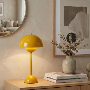 Könnte beinhalten: Eine leuchtend gelbe Tischlampe mit einem pilzförmigen Lampenschirm. Die Lampe hat einen schlanken Stiel und einen runden Sockel. Sie steht auf einer Holzoberfläche, mit Büchern und einer Vase in der Nähe. Eine dekorative Leuchte.