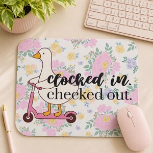 May include: A rectangular mousepad with a floral pattern and a cartoon goose riding a pink scooter. The mousepad has the text "clocked in. checked out." in black script. A pink mouse and keyboard are visible.