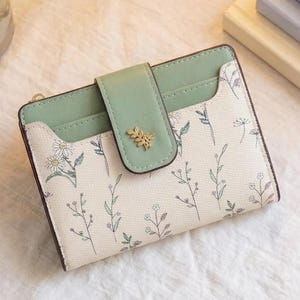 May include: A small, bi-fold wallet with a light green flap and a cream-colored body. The wallet features a delicate floral pattern in light green and a gold-colored leaf emblem on the flap. The wallet is closed.