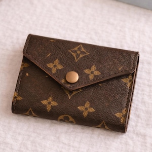 May include: A brown wallet with a flap closure and a gold-colored snap. The wallet features a repeating pattern of interlocking floral and geometric shapes. The wallet is made of a textured material.
