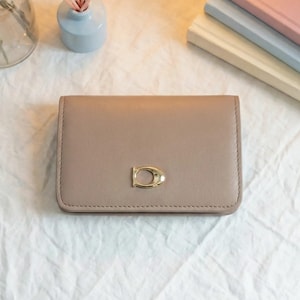 Puede incluir: Un tarjetero rectangular de cuero color topo con un emblema de logotipo en forma de D de metal dorado centrado en la solapa frontal. El accesorio tiene costuras tonales visibles a lo largo de los bordes y un acabado mate liso.