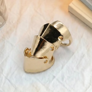 May include: A gold-toned metal full-finger ring featuring three articulated, overlapping segments. Each segment has a smooth, polished surface with a small circular cutout on the side, designed to cover the length of a finger with a hinged structure.