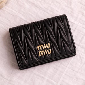 May include: Black quilted leather wallet with a geometric pattern. The wallet features a gold-colored "MIU MIU" logo on the front. The wallet is rectangular and appears to be closed. The background is a white, textured surface.