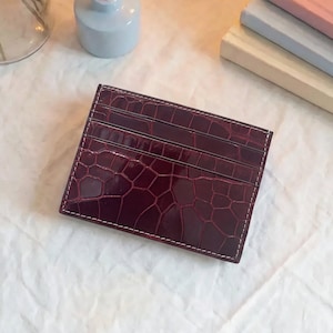May include: A rectangular card holder in a deep burgundy colour with a glossy finish and a textured crocodile skin pattern. The accessory features three horizontal slots for cards and is constructed with visible white stitching along the edges.