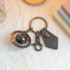 Puede incluir: Un llavero de metal oscuro que presenta un dije de orbe esférico rodeado por un anillo, una anilla circular, un mosquetón y una etiqueta de cuero gris oscuro con un pequeño emblema metálico circular con un diseño de logotipo planetario.