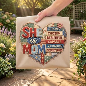 May include: A beige fabric square featuring a heart-shaped design. The heart is crafted from patchwork fabric and displays the words "SHE is MOM" in red and blue. The heart also includes the words "STRONG, CHOSEN, BEAUTIFUL, CAPABLE, VICTORIOUS, GREATLY LOVED". A small pink flower is attached.