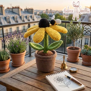 May include: A handcrafted crochet sunflower with yellow petals, a black center, and a bee, sits in a brown crochet pot. The flower is on a wooden table with a drawing of a sunflower, a pen, and a compass.