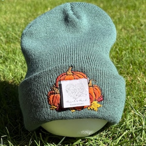 May include: A sage green knit beanie featuring an embroidered design of orange pumpkins and yellow leaves. A white square fabric label is stitched over the center of the embroidery, displaying a floral heart graphic and the text Heart & Hand EMBROIDERY.