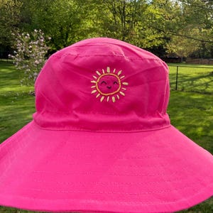 May include: A bright pink bucket hat featuring a small, embroidered yellow sun design on the front. The sun has a simple, smiling face with closed eyes and a small mouth, stitched in black thread. The hat has a wide, downward-sloping brim.
