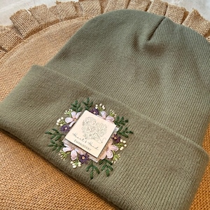 May include: An olive green ribbed knit beanie featuring a square white patch with the text Heart & Hand EMBROIDERY. The patch is surrounded by detailed floral embroidery in shades of purple, light pink, white, and dark green thread.