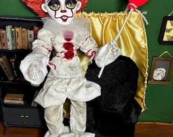May include: A handmade doll of a clown with red hair, white ruffled clothing, and red shoes. The doll has a painted face with yellow eyes and a red smile. It holds a red balloon on a string. The background is green with a gold curtain.