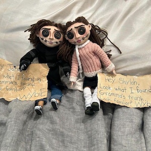 May include: Two handmade dolls with button eyes and stitched smiles, dressed in miniature clothing. One doll wears a black coat and jeans, the other a pink sweater and black skirt. Each doll holds a sign with handwritten text.