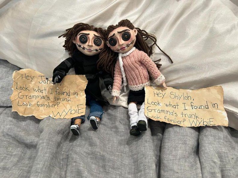 May include: Two handmade dolls with button eyes and stitched smiles, dressed in miniature clothing. One doll wears a black coat and jeans, the other a pink sweater and black skirt. Each doll holds a sign with handwritten text.