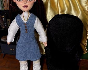 May include: A handmade doll with long brown hair, red hair ties, and a denim blue dress over a white long-sleeved shirt. The doll has a gold necklace and black shoes. A black object is behind the doll.