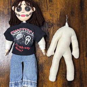 May include: A handmade doll with button eyes, stitched mouth, and dark brown yarn hair. The doll wears a black t-shirt with a graphic and blue jeans. A separate, unadorned doll body is also present.