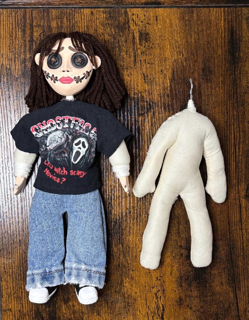 May include: A handmade doll with button eyes, stitched mouth, and dark brown yarn hair. The doll wears a black t-shirt with a graphic and blue jeans. A separate, unadorned doll body is also present.