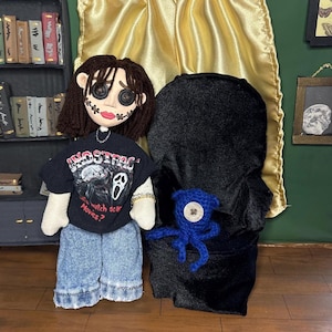 May include: Handmade doll wearing a black t-shirt with the text "Ghostface Movies?" and denim shorts. The doll has button eyes and a stitched face. A black hooded figure and a blue crocheted octopus are also present.