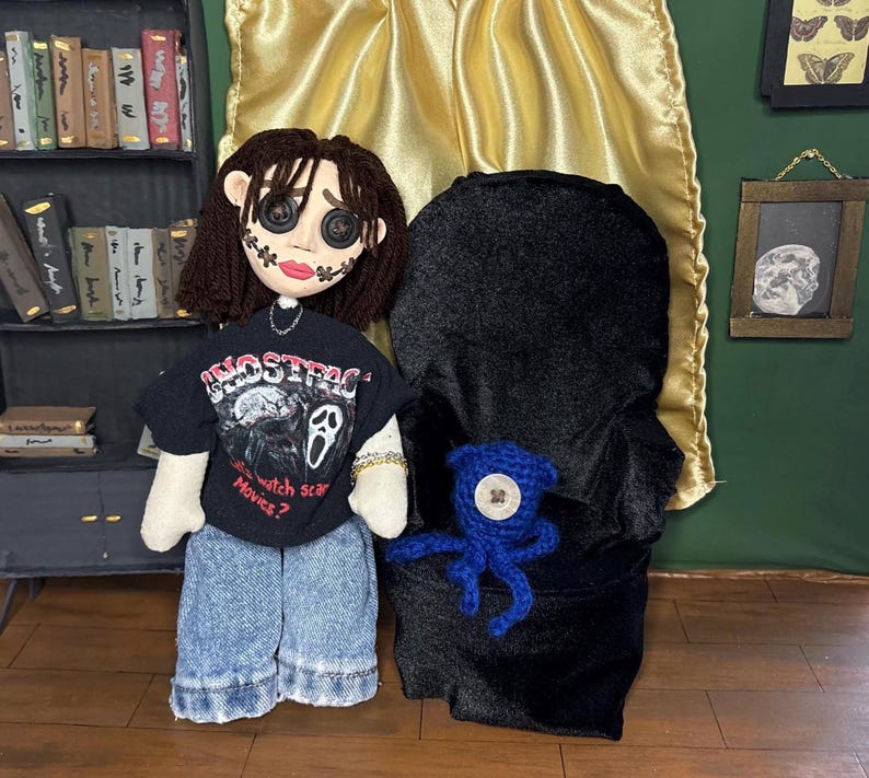 May include: Handmade doll wearing a black t-shirt with the text "Ghostface Movies?" and denim shorts. The doll has button eyes and a stitched face. A black hooded figure and a blue crocheted octopus are also present.