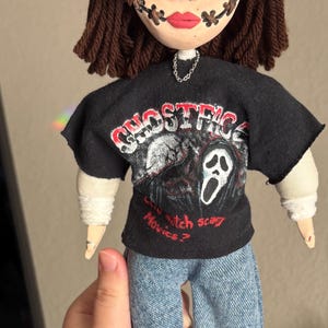 May include: A handmade doll with a button-eyed face, stitched mouth, and brown yarn hair. The doll wears a black t-shirt with a graphic of a ghost face and the text "Ghostface". It is dressed in blue jeans.
