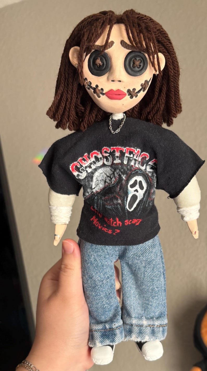 May include: A handmade doll with a button-eyed face, stitched mouth, and brown yarn hair. The doll wears a black t-shirt with a graphic of a ghost face and the text "Ghostface". It is dressed in blue jeans.
