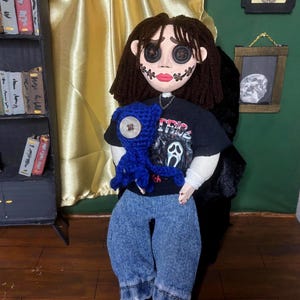 May include: A doll with button eyes, stitched mouth, and dark hair, wearing a black t-shirt with a graphic print and blue jeans. The doll is holding a blue crocheted creature. The background includes a bookshelf and a framed picture.