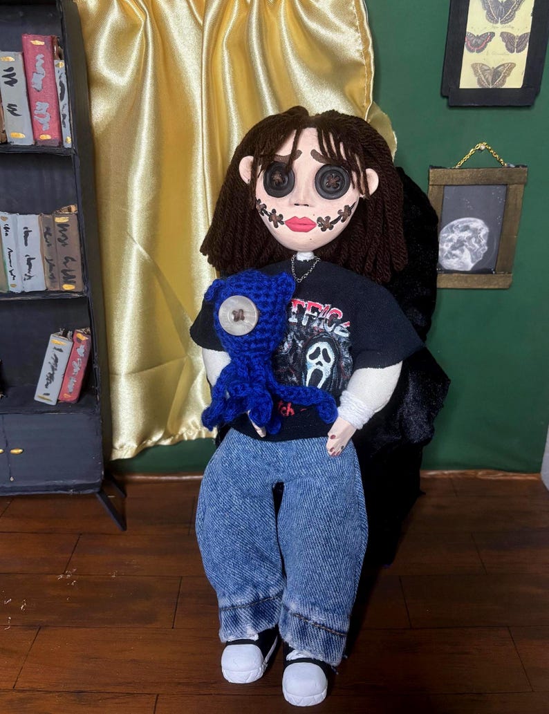 May include: A doll with button eyes, stitched mouth, and dark hair, wearing a black t-shirt with a graphic print and blue jeans. The doll is holding a blue crocheted creature. The background includes a bookshelf and a framed picture.