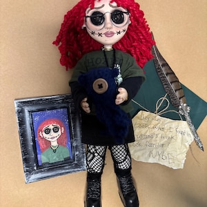 May include: A handmade doll with red yarn hair, round black glasses, and stitched facial features. The doll wears a green shirt, black skirt, and boots, holding a blue stuffed animal. A framed picture and a note with text are also present.