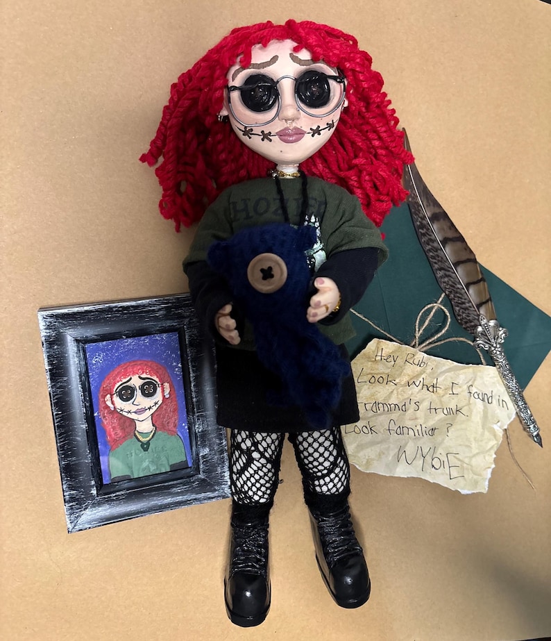 May include: A handmade doll with red yarn hair, round black glasses, and stitched facial features. The doll wears a green shirt, black skirt, and boots, holding a blue stuffed animal. A framed picture and a note with text are also present.