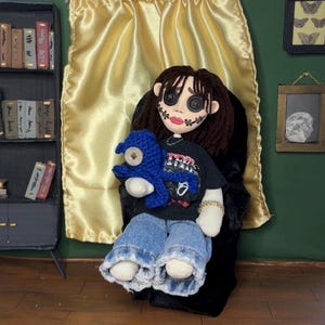 May include: A handmade doll with button eyes, stitched facial features, and brown yarn hair. The doll wears a black t-shirt, blue jeans, and holds a blue crocheted creature. The doll is seated in a black chair against a gold curtain.