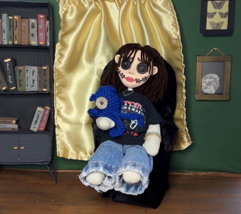 May include: A handmade doll with button eyes, stitched facial features, and brown yarn hair. The doll wears a black t-shirt, blue jeans, and holds a blue crocheted creature. The doll is seated in a black chair against a gold curtain.