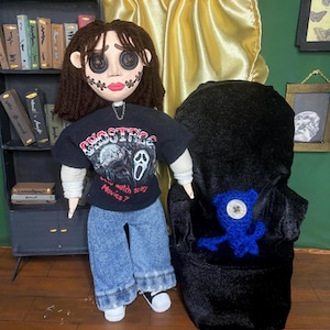 May include: A doll with button eyes, stitched mouth, and brown hair, wearing a black t-shirt with a Scream movie graphic and the text "...watch scary Movies?". The doll is wearing blue jeans and white sneakers. A blue crocheted creature sits in a black chair.
